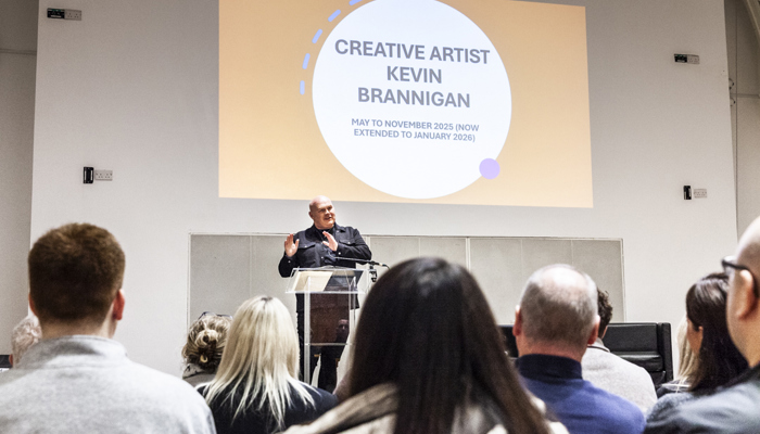 A man speaks on stage under a screen displaying "Creative Artist Kevin Brannigan".