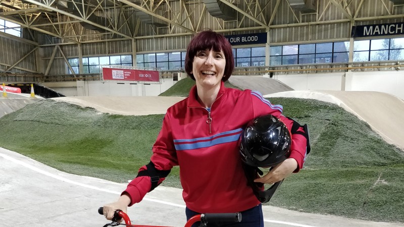 A person standing smiling and standing over a BMX bike. There is a BMX track in the background.