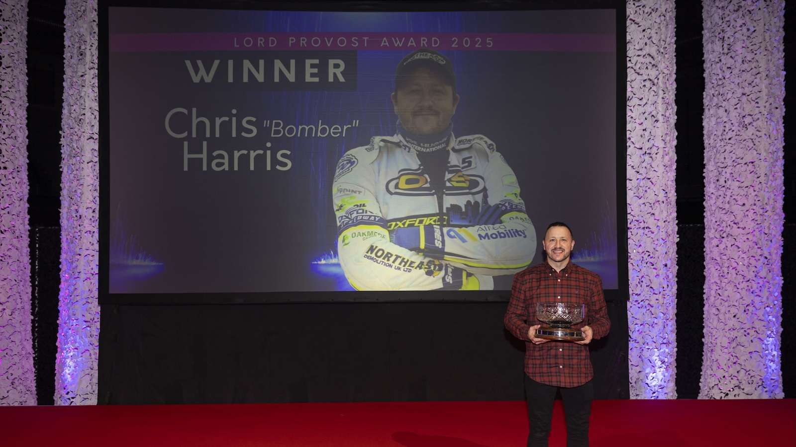 A man wearing a red shirt holding an award stands in front of a large screen which displays the words 'Lord Provost Award 2025 winner' and 'Chris Bomber Harris'
